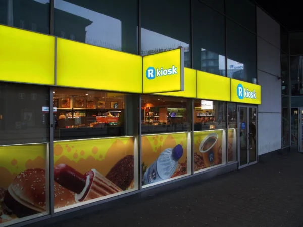 R-kiosk valguskastid kollase fassaadipaneeliga kioski sissepääsu juures õhtul