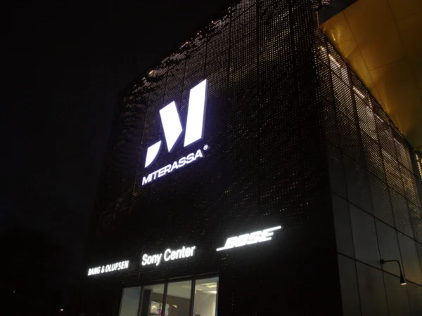 Miterassa valgustatud M-logo ja tekst perforeeritud musta fassaadipaneeli pinnal
